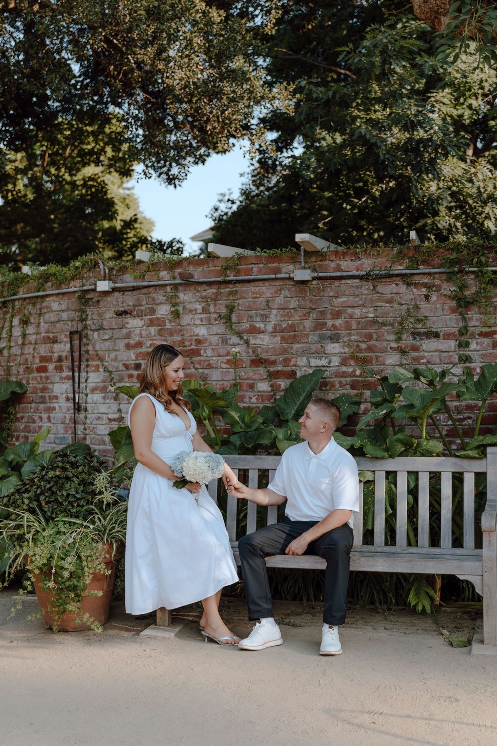 Two people in white outfits share a moment on a garden bench against a rustic brick wall with greenery.