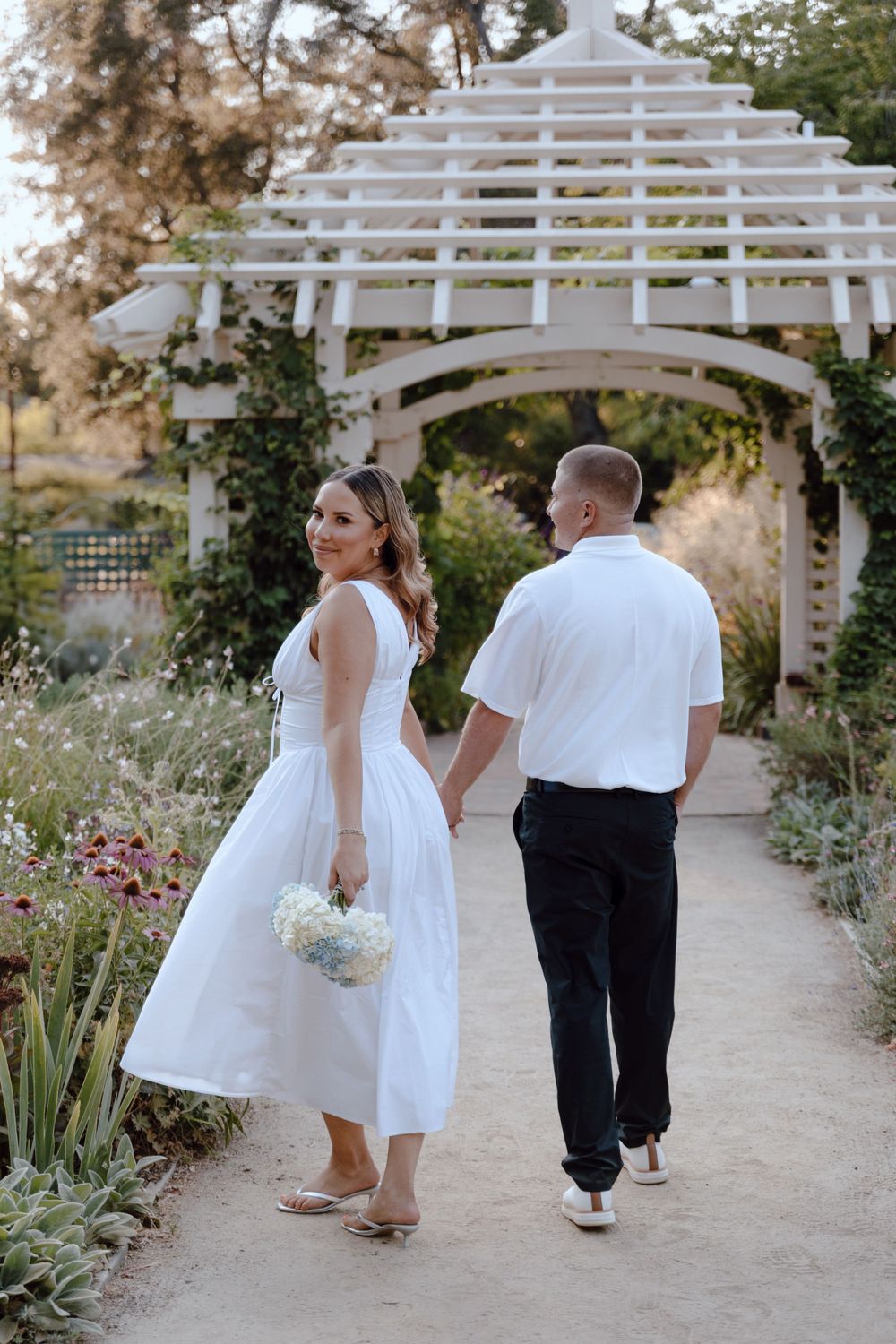 A couple walking on a garden path under a white pergola with flowers and greenery around them.