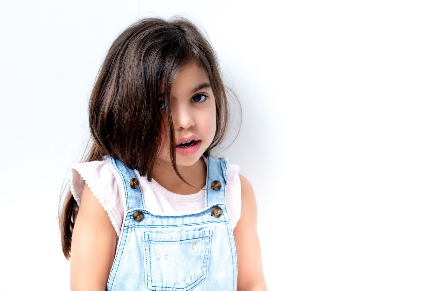 Young child wearing light denim overalls and a white shirt poses against white background.