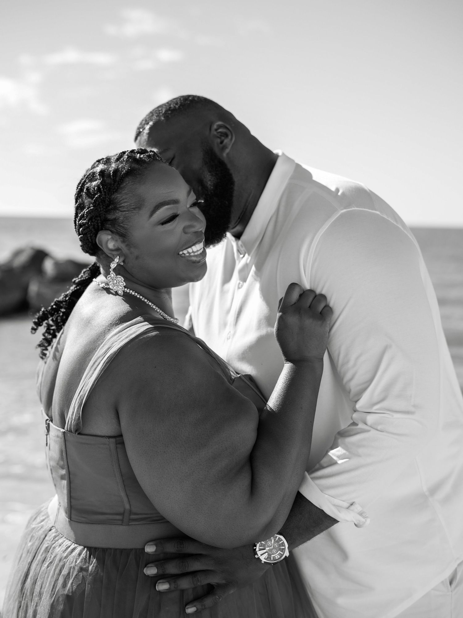 A romantic black and white beachside photo of a couple embracing and sharing a tender moment together.