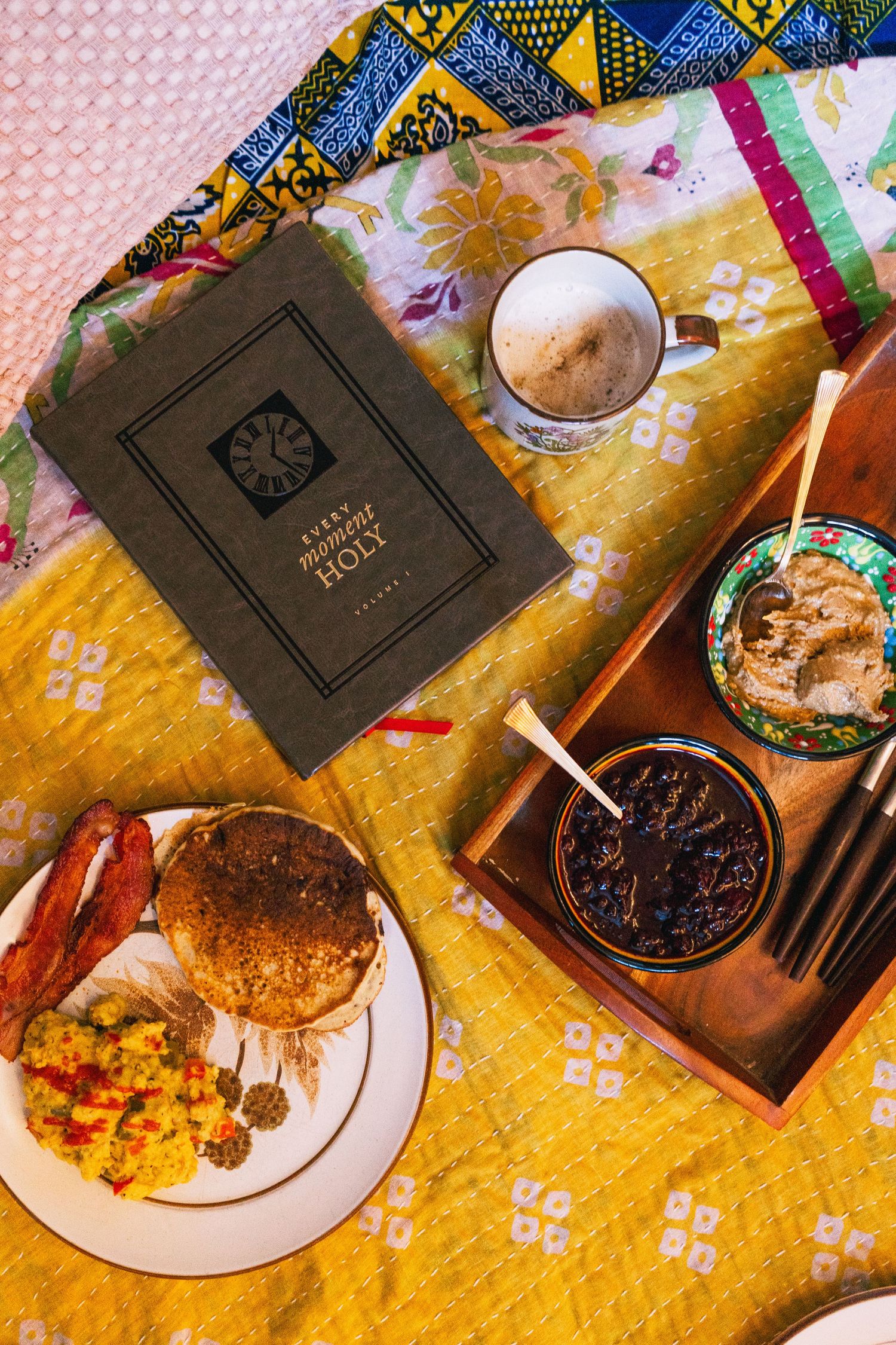 A black hardcover book rests on a yellow floral tablecloth alongside plated food, drinks and candlesticks for a cozy meal.