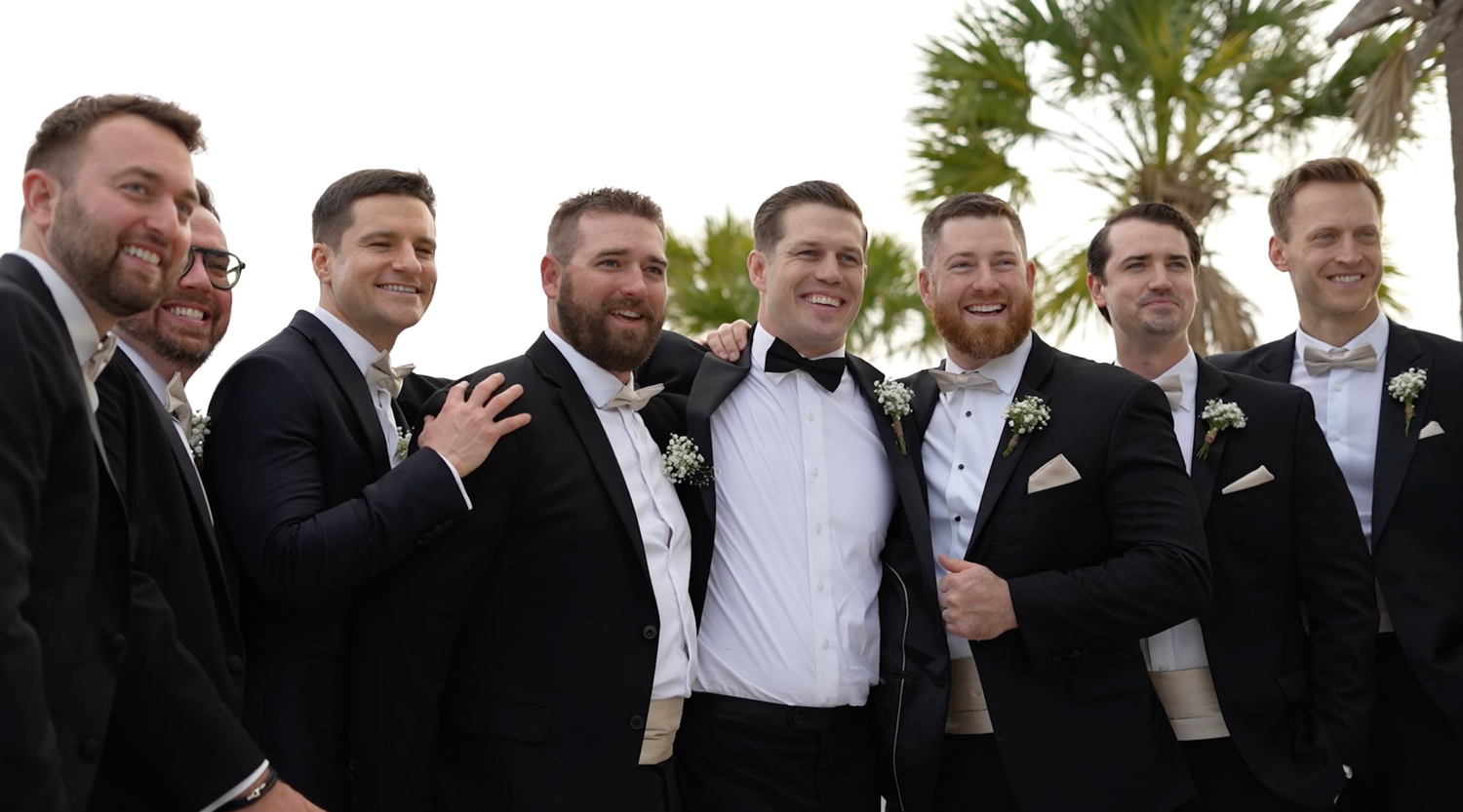 A group of groomsmen in black tuxedos laughing and smiling together outdoors with palm trees in the background.