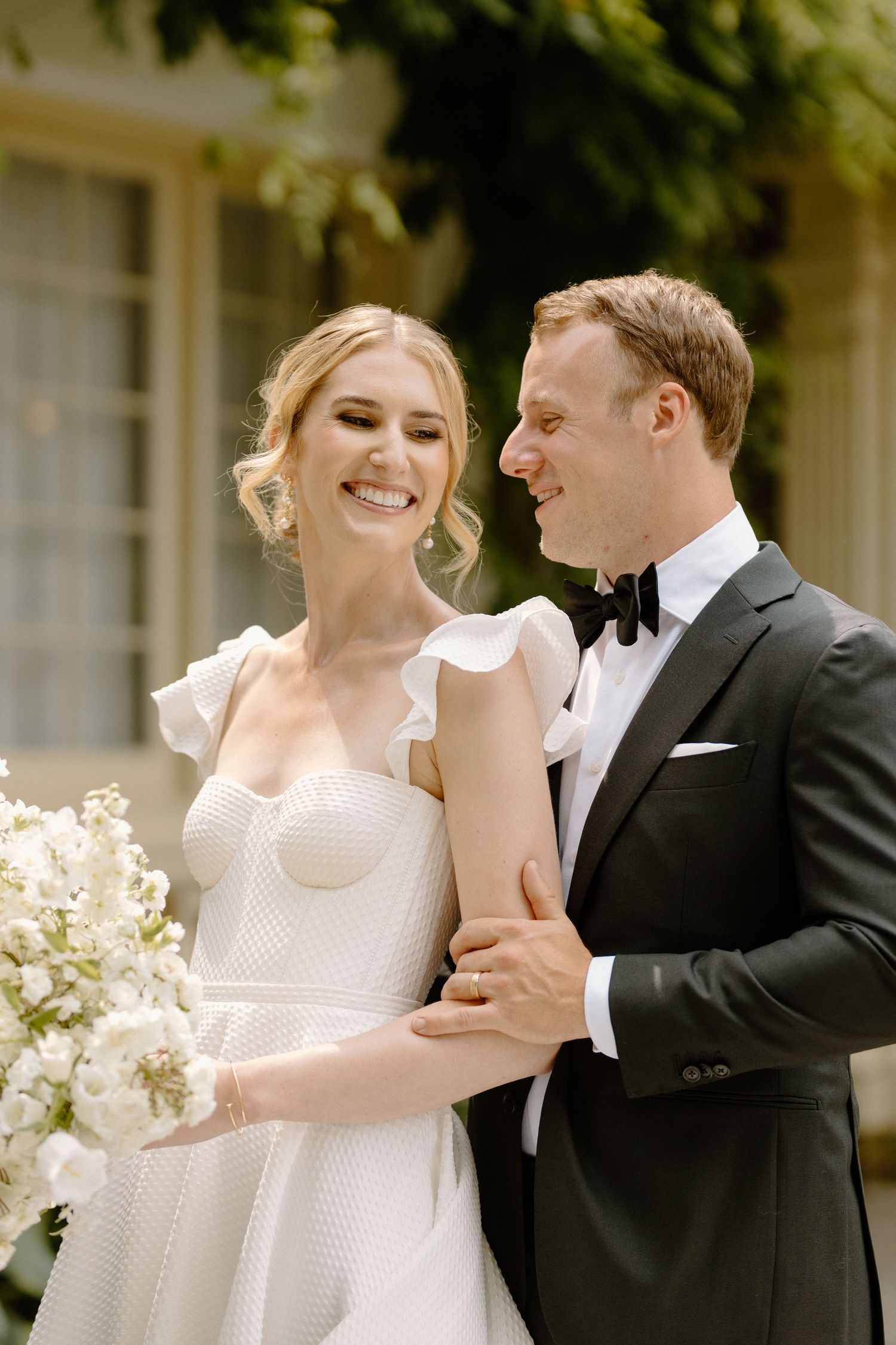 Elegant bride and groom share a happy moment together outdoors with delicate white floral arrangements in the background.