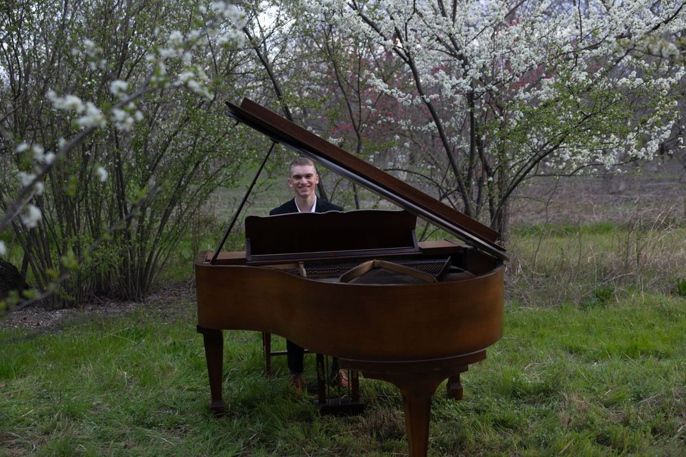 A grand piano sits outdoors in a garden setting with blooming cherry blossoms and spring greenery around it.