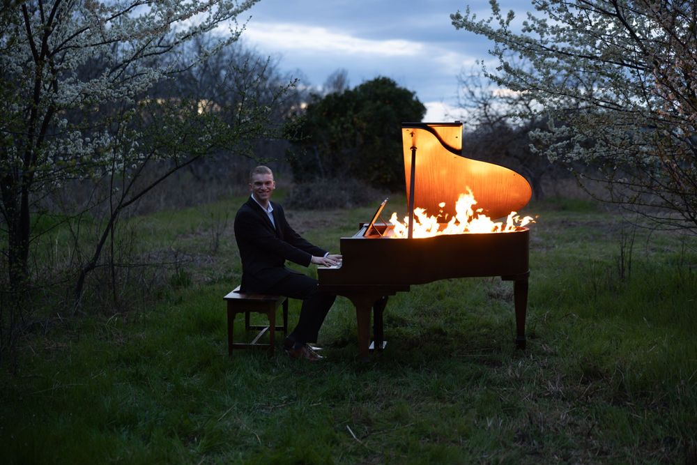 Grand piano engulfed in flames illuminates a dark outdoor garden setting during twilight.