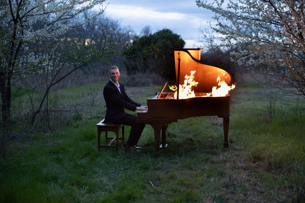 An illuminated grand piano glows orange in a dusky outdoor setting with trees and grass in the background.