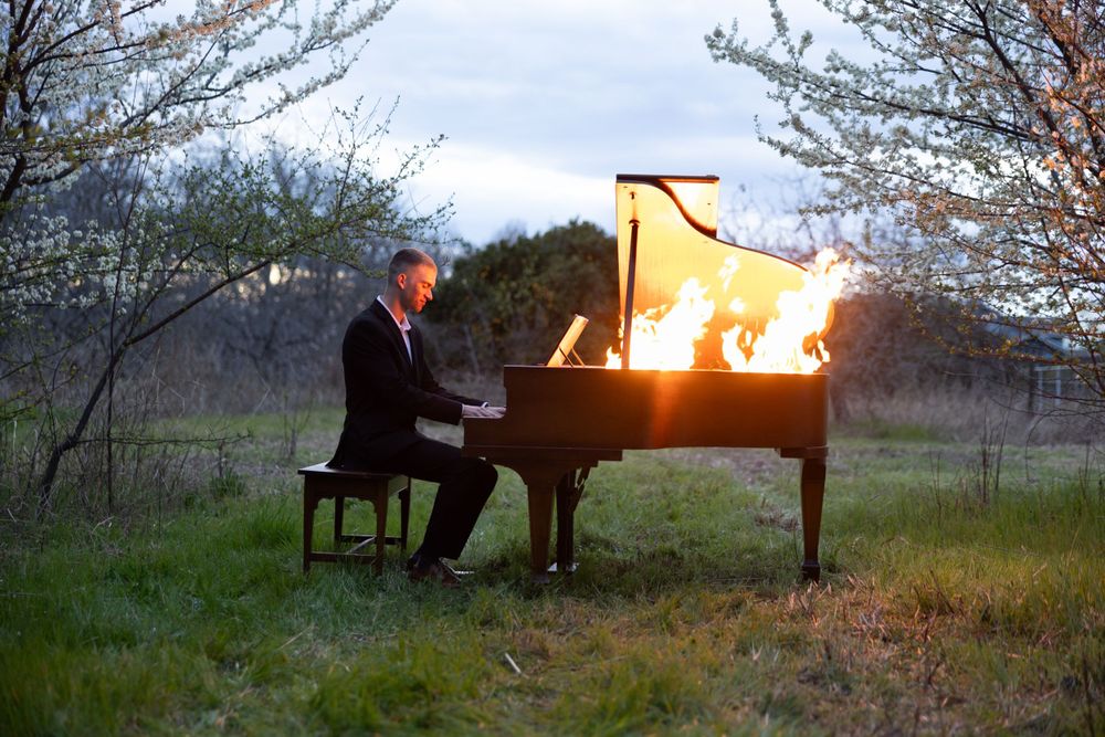 Dramatic scene of musician playing piano engulfed in flames during outdoor twilight performance.