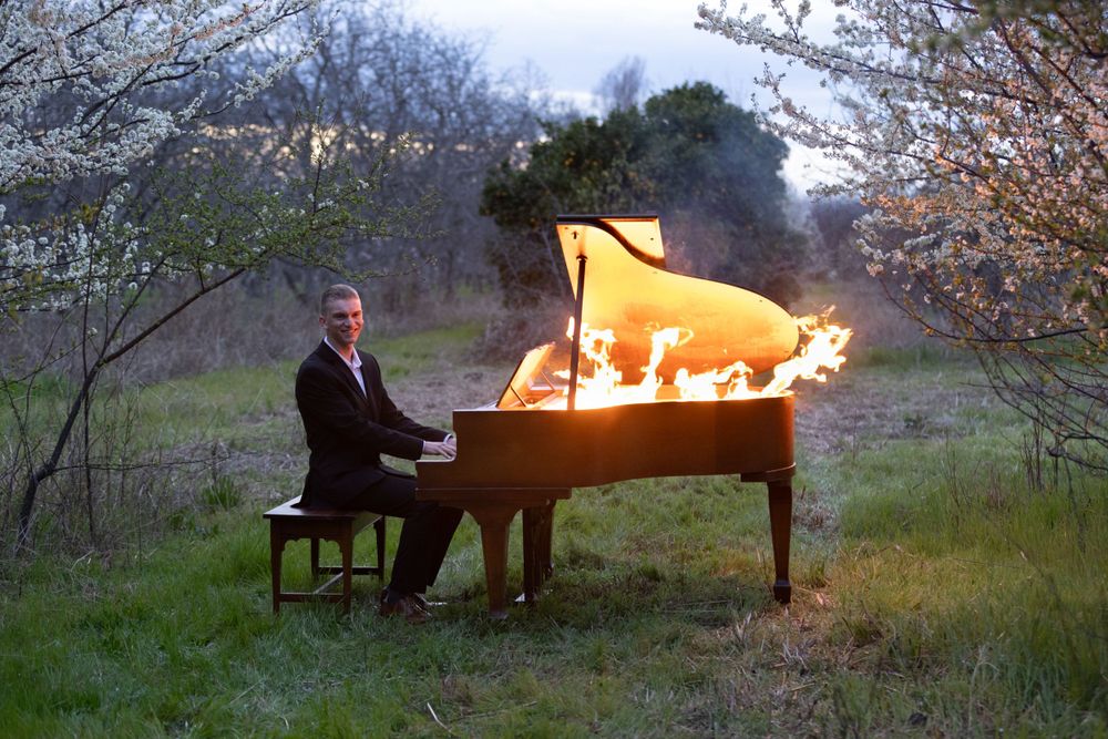 A grand piano engulfed in flames sits in a meadow at dusk while being played.