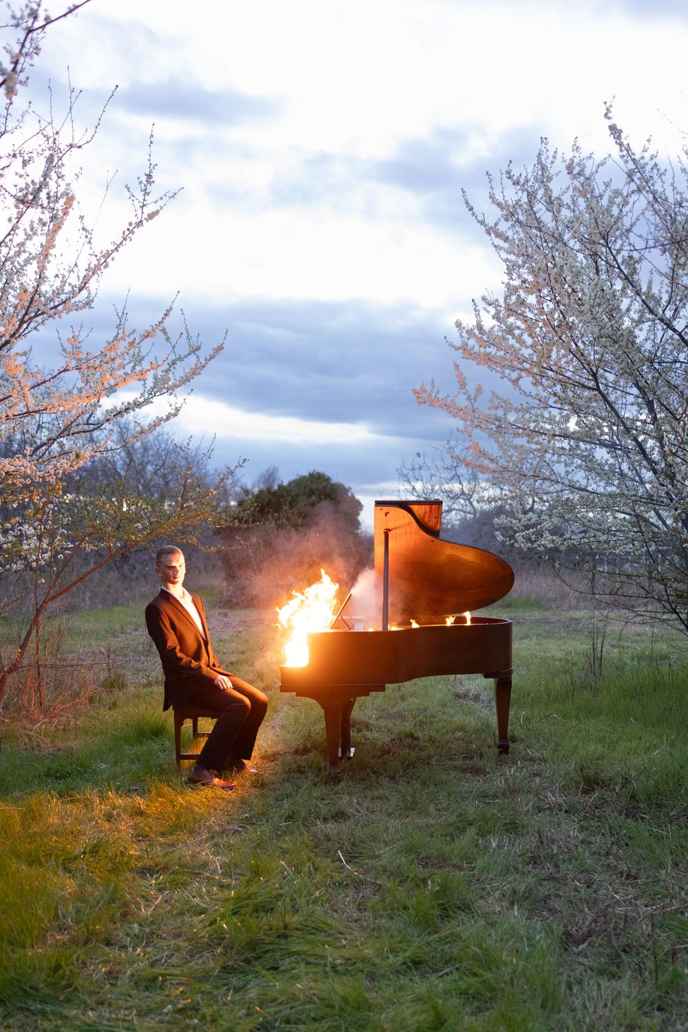 A dramatic scene shows a burning grand piano in a field with spring blossoms and cloudy skies.