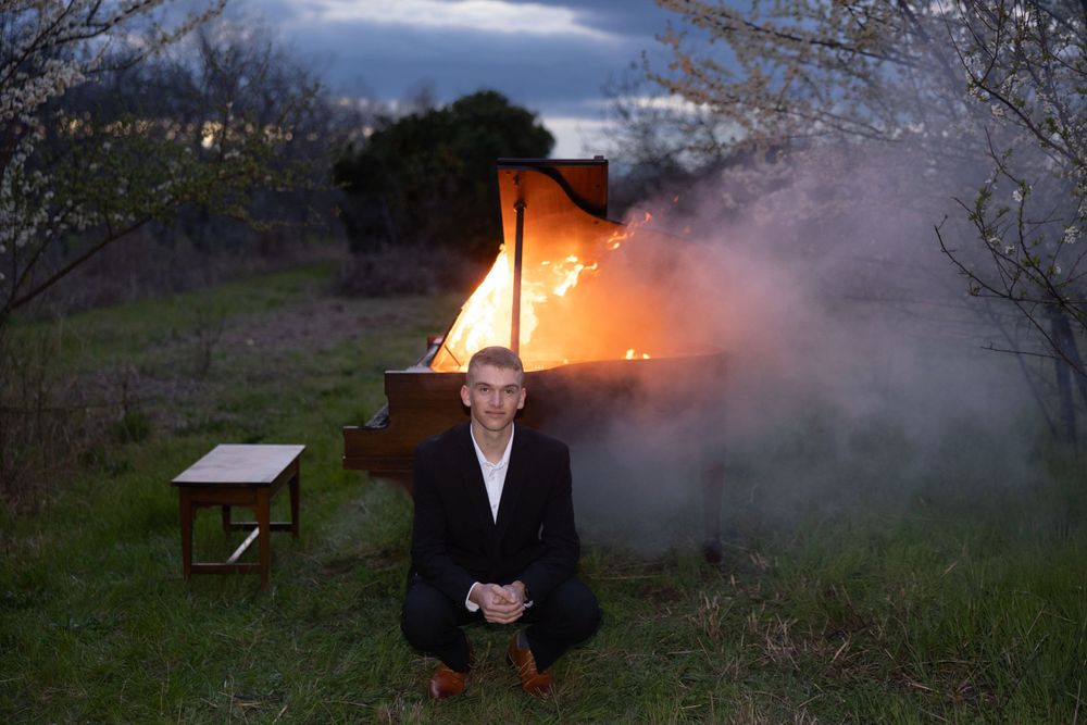 Figure in dark suit crouches near burning piano outdoors at dusk with smoke billowing in background.