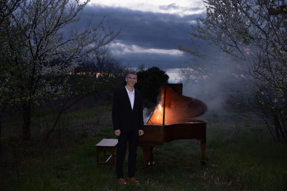 A grand piano stands illuminated in a dark field at dusk with dramatic storm clouds overhead.