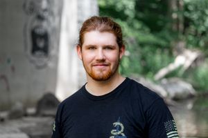 Portrait of person in black t-shirt with beard against blurred outdoor background.