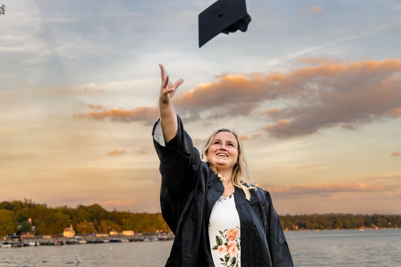 Graduate celebrates by tossing cap in the air against sunset sky with lake in background.