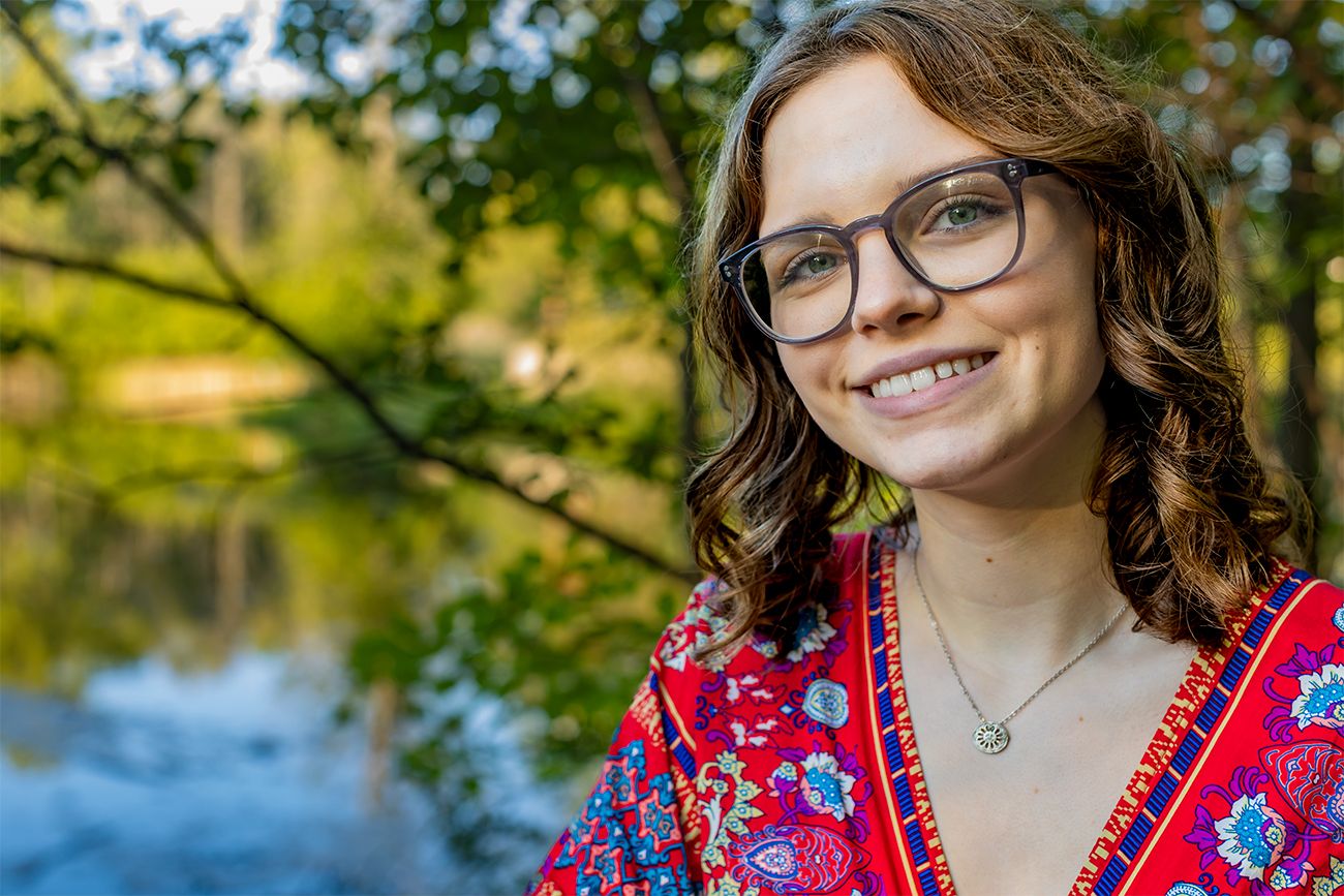Portrait of someone wearing glasses and a red floral top smiling outdoors near a lake with trees in background.