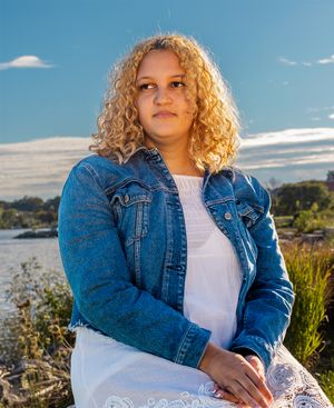 A person in a denim jacket and white top poses outdoors with curly blonde hair against a scenic coastal backdrop.
