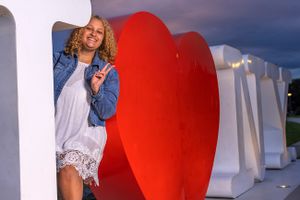 Woman in denim jacket beside large red heart sculpture against white wall at dusk.