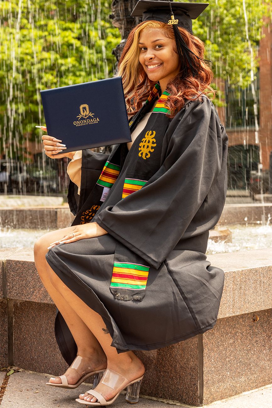 Graduate wearing black academic regalia with colorful kente stole displays diploma while sitting outdoors.