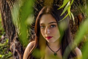 Portrait of a young woman with dark hair peering through green leaves in natural lighting.