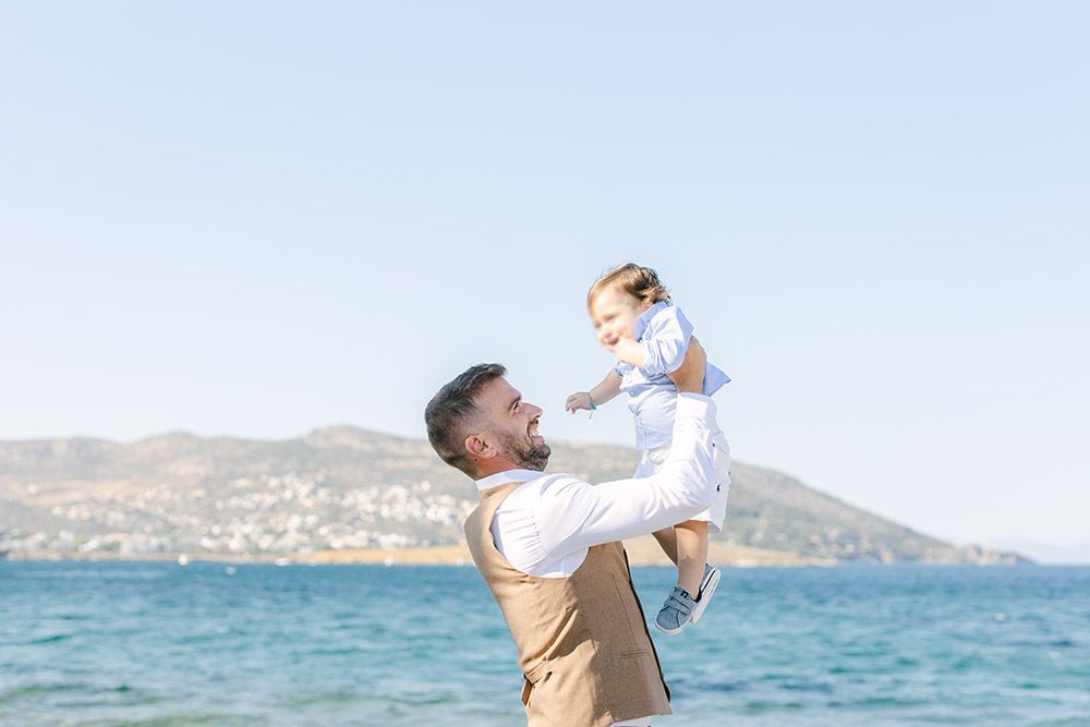 Parent and baby playing together on a sunny beach with blue ocean water and mountains in the background.