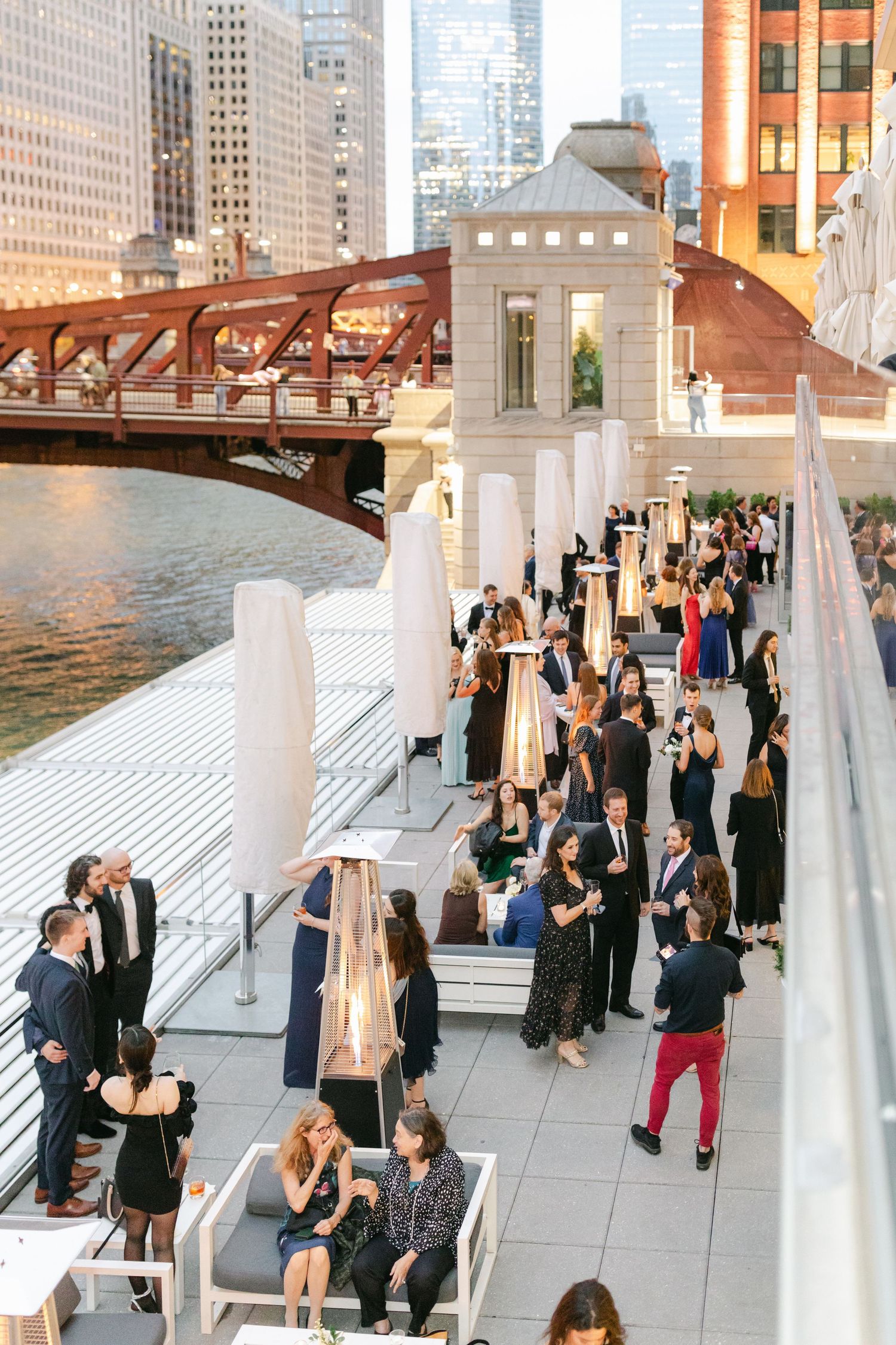 People gather at an outdoor riverside venue in downtown Chicago with the iconic Lake Street Bridge visible in the background.