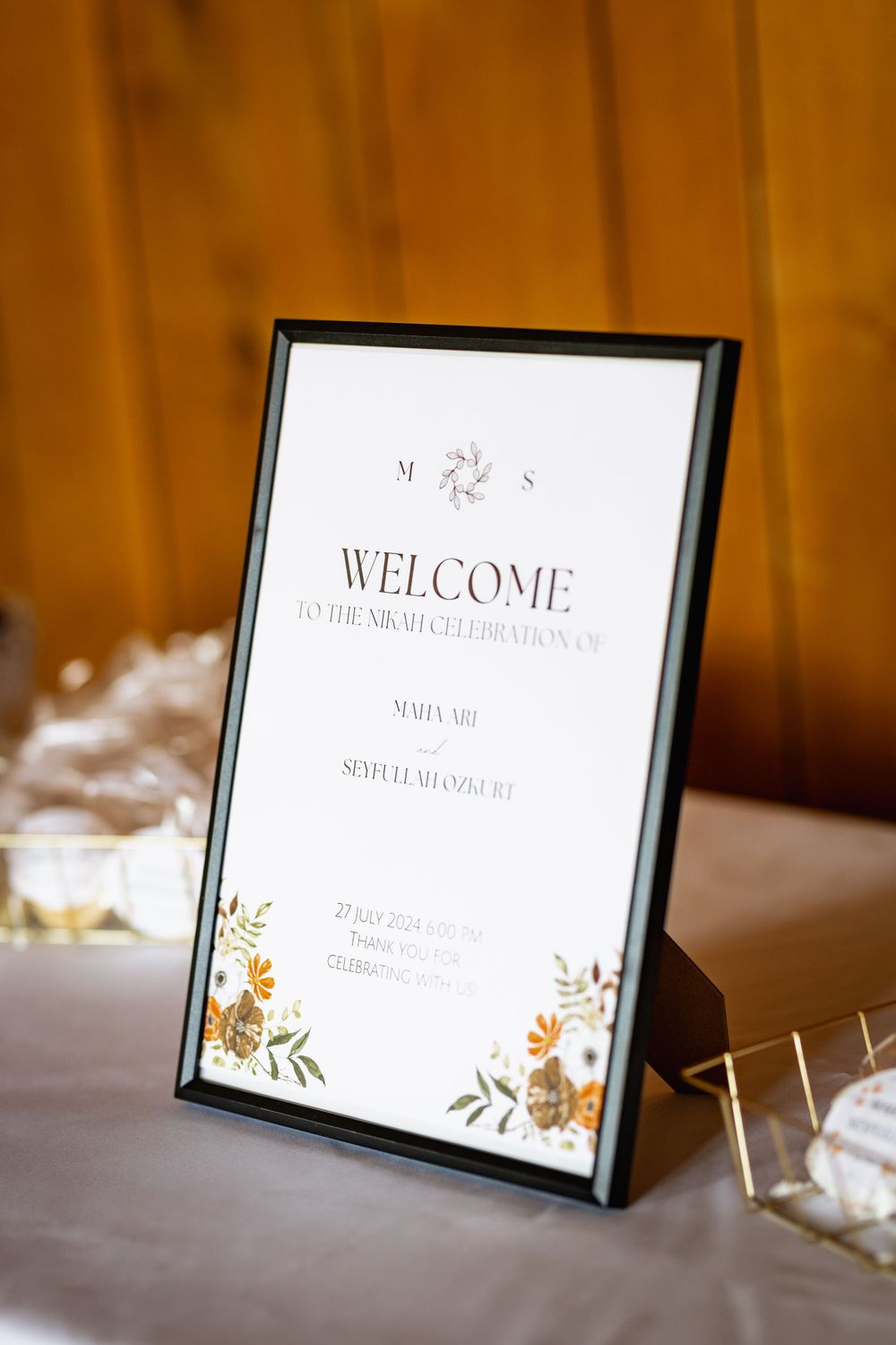 A framed wedding welcome sign with floral details sits on a white tablecloth against an orange backdrop.