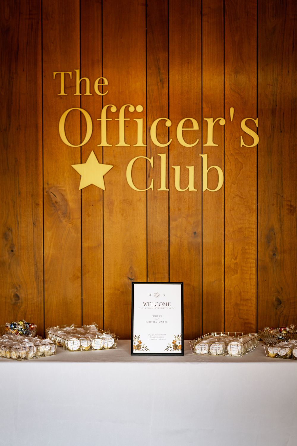 Gold lettered "The Officer's Club" sign with star emblem mounted on wooden paneled wall above small decorative items.