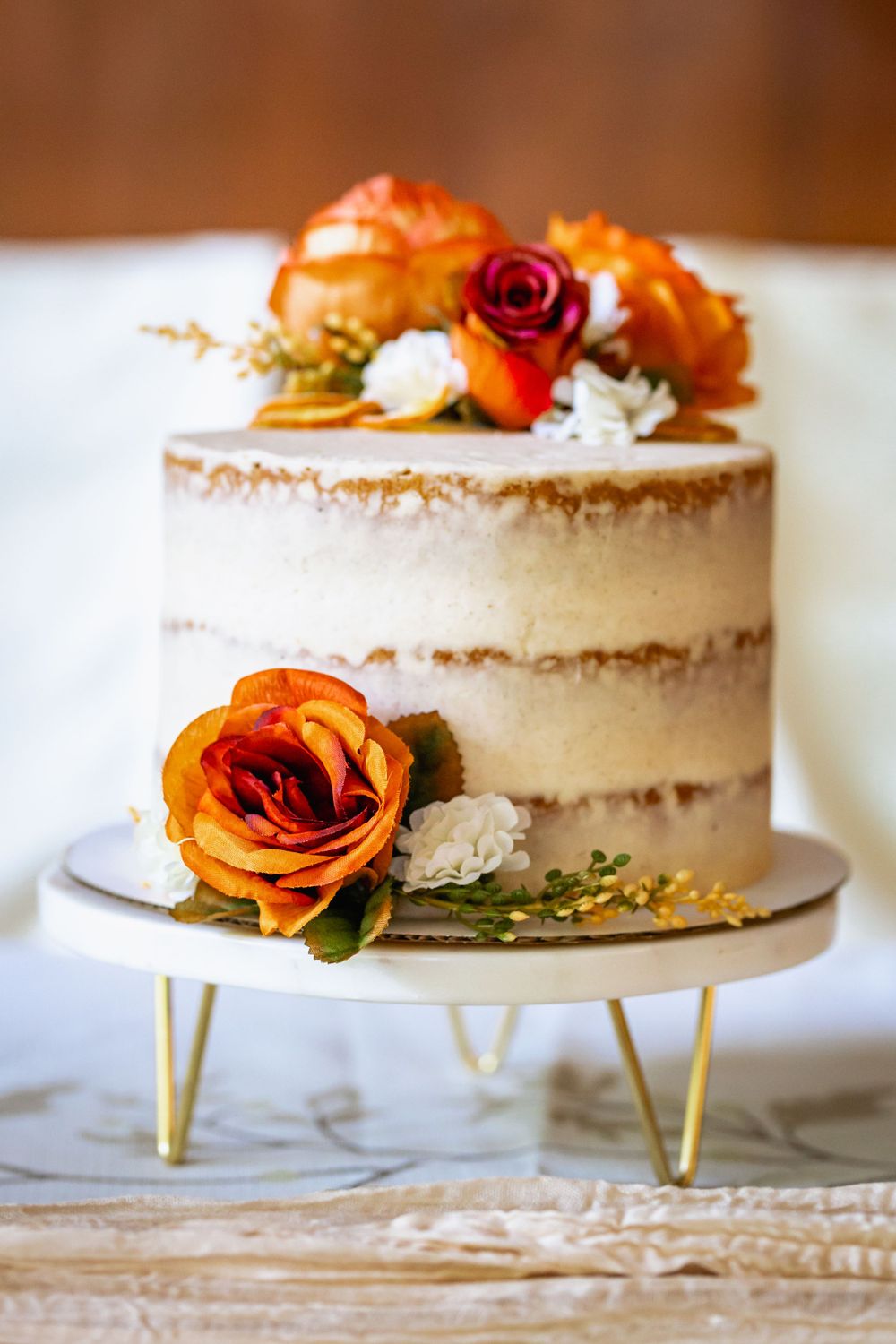 A semi-naked wedding cake decorated with orange and burgundy roses on a gold-footed cake stand.