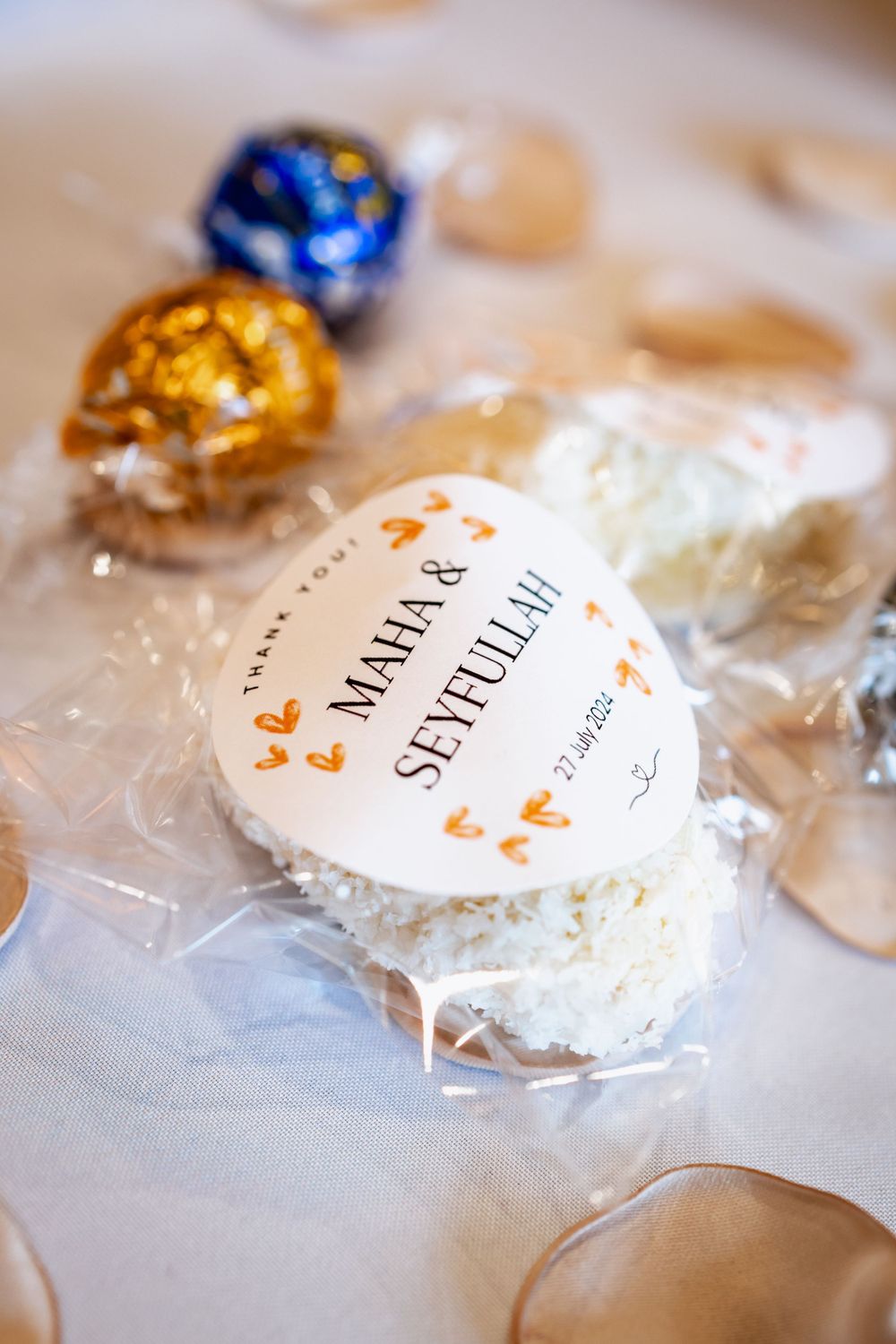 Custom wedding favor with name tag showing Maha & Septuhar surrounded by gold and blue wrapped treats on white tablecloth.