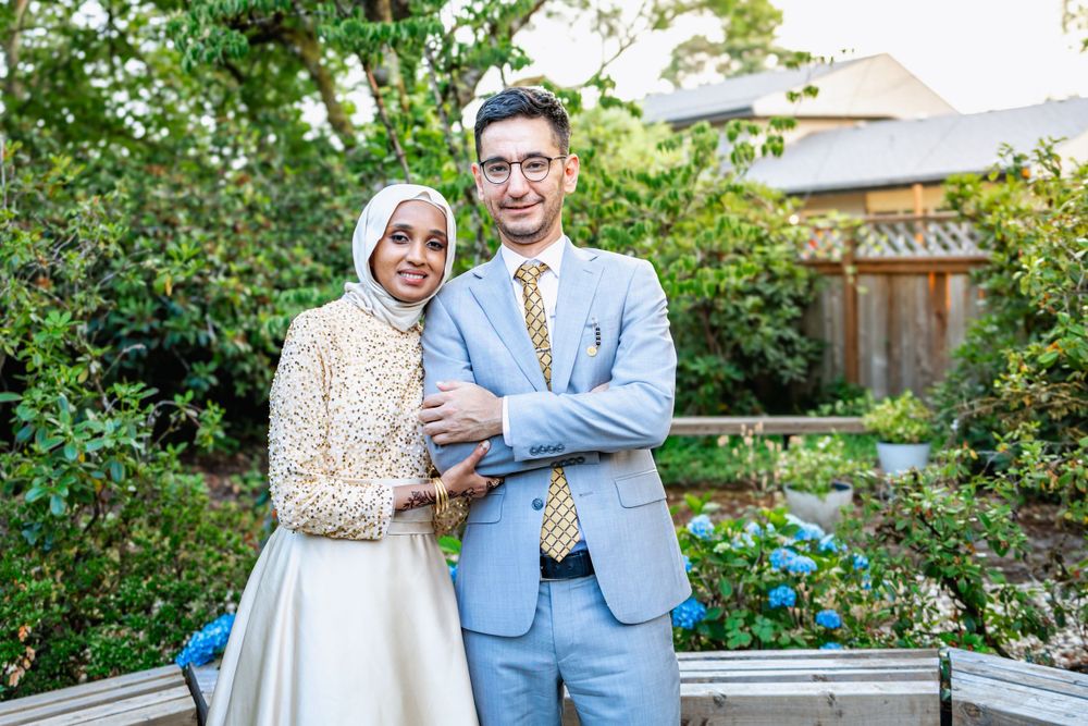 A couple stands together in a lush garden setting, wearing formal attire with a light blue suit and cream-colored dress.