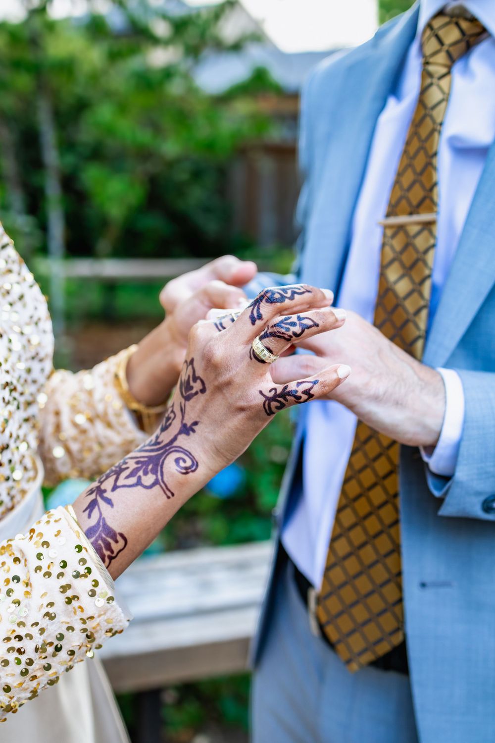 Close-up of hands adorned with henna designs during a wedding ceremony, with blue suit jacket and patterned tie visible.