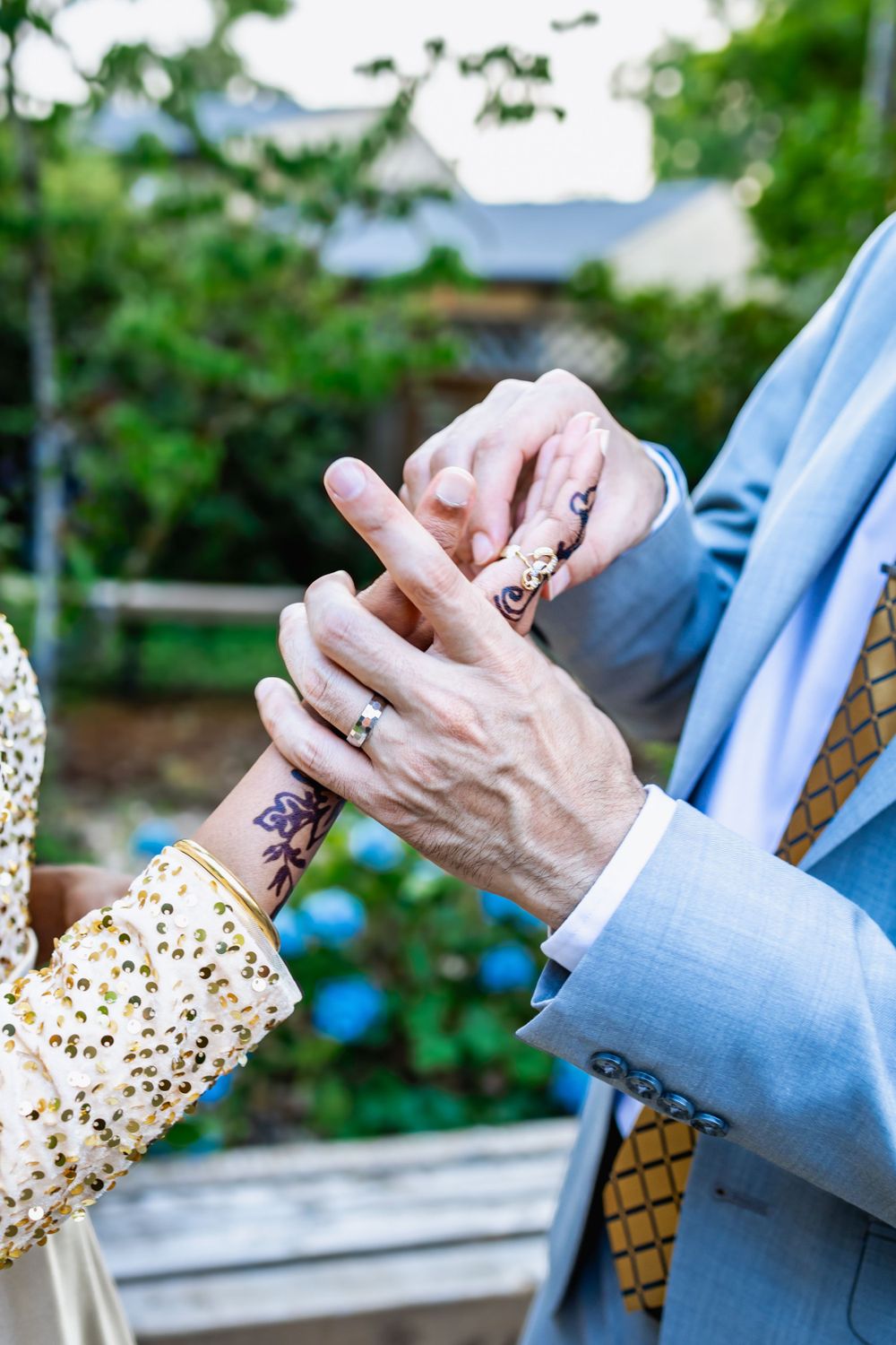 Detail shot of hands with intricate henna designs exchanging rings during a wedding ceremony against green foliage.