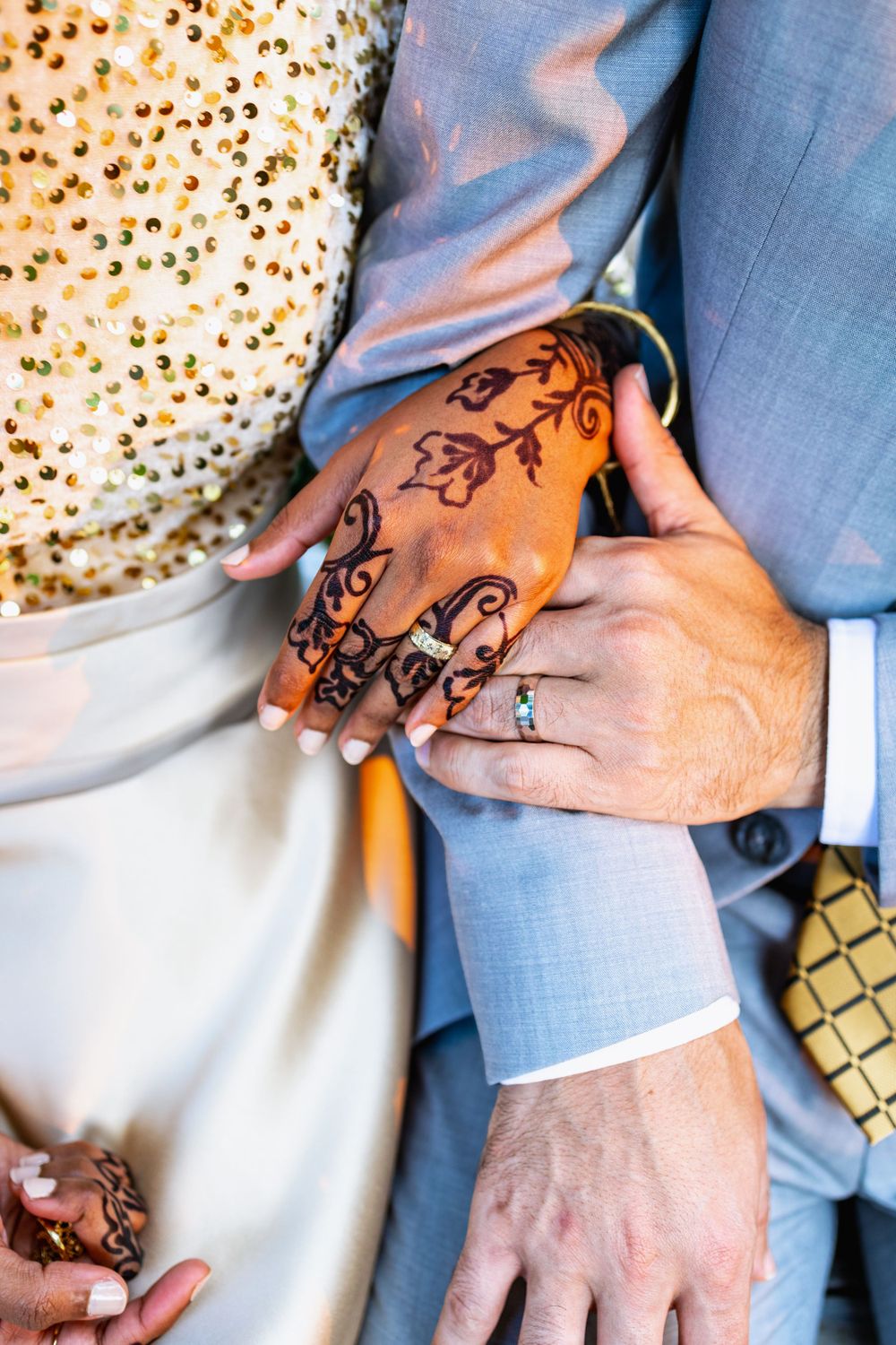 Close-up detail of hands with intricate henna tattoo design and wedding rings against formal attire.