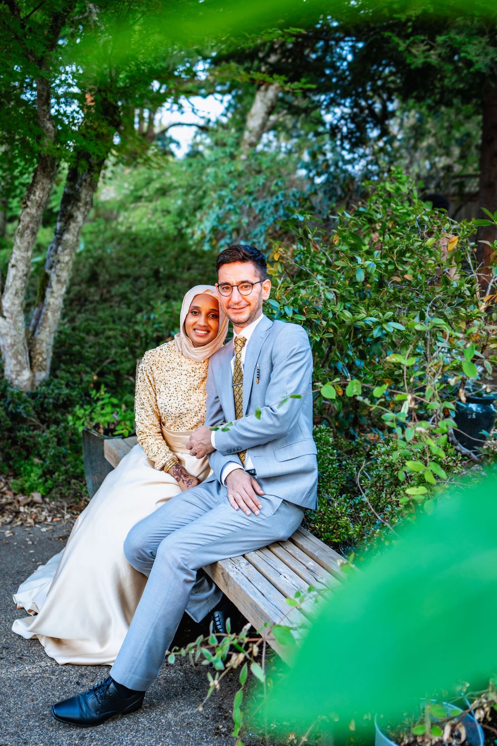 A couple sits together on a wooden bench surrounded by lush green garden foliage.