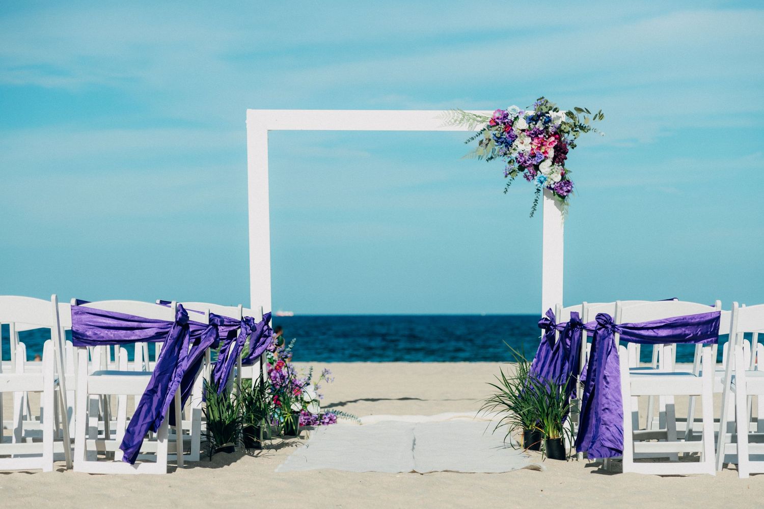 Beach wedding arch decorated with purple ribbons and flowers against ocean backdrop.