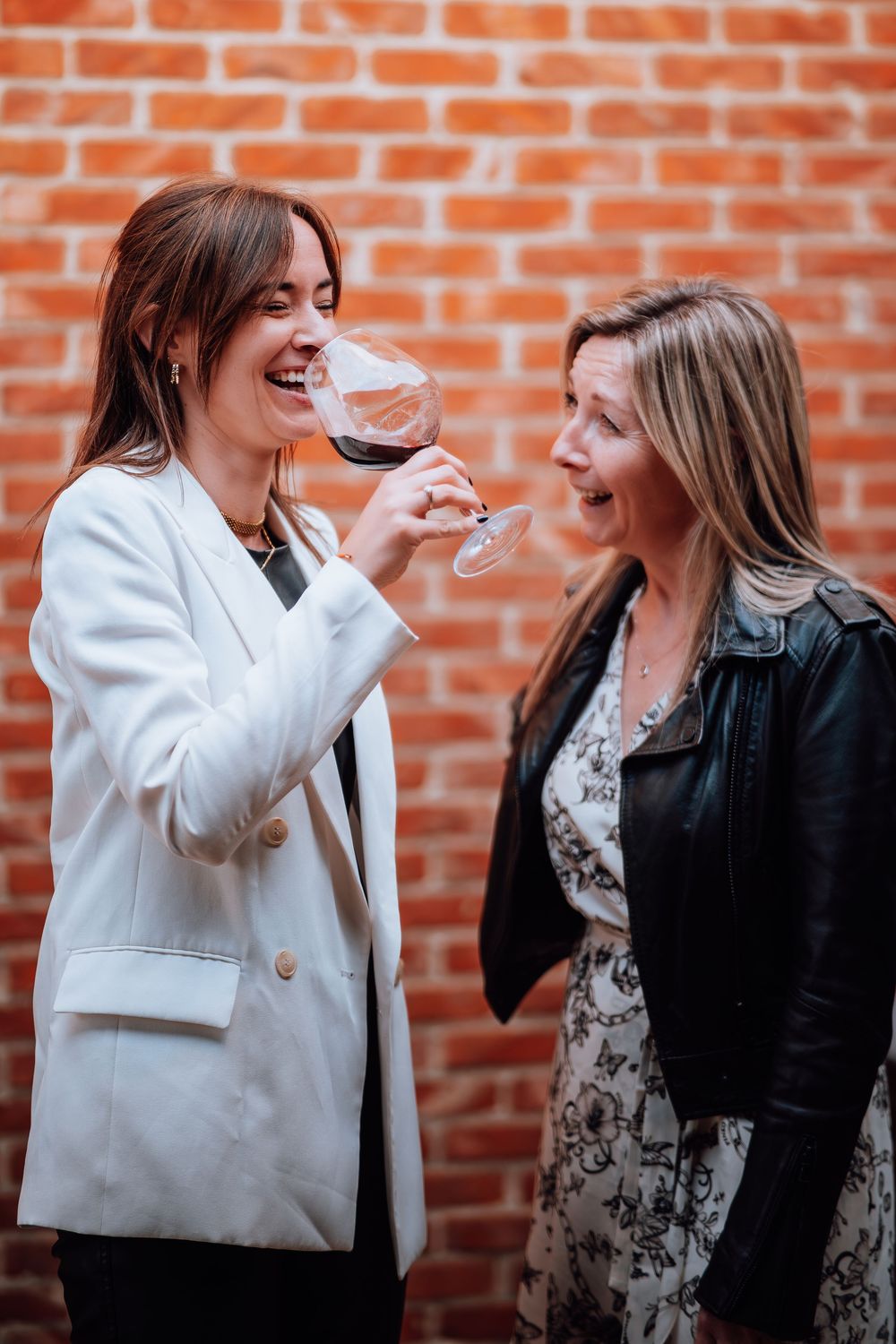 Twee vrienden genieten van rode wijn en lachen samen tegen een bakstenen muur op de achtergrond.