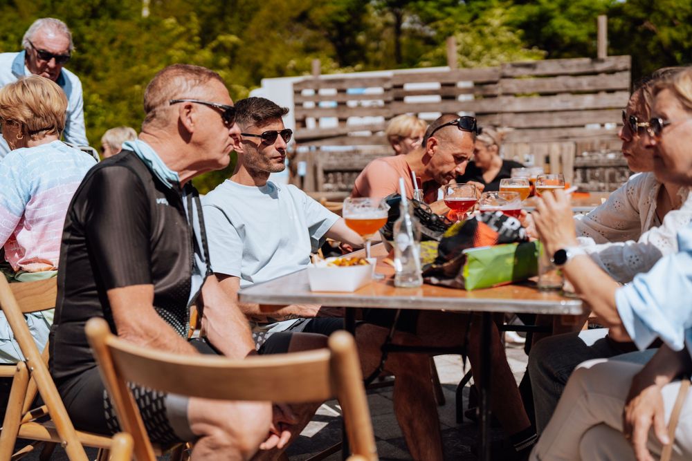 Een groep wielrenners geniet op een zonnige dag van een maaltijd in de buitenlucht aan een houten picknicktafel.