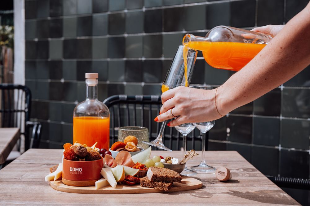 Iemand schenkt een feloranje cocktail in op een houten tafel met een charcuterieplank en hapjes.