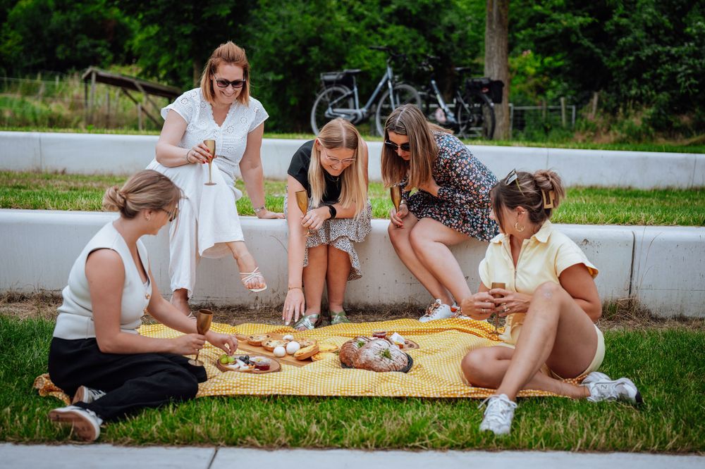 People sitting on blankets enjoying an outdoor picnic with food and drinks on a grassy area near a path.