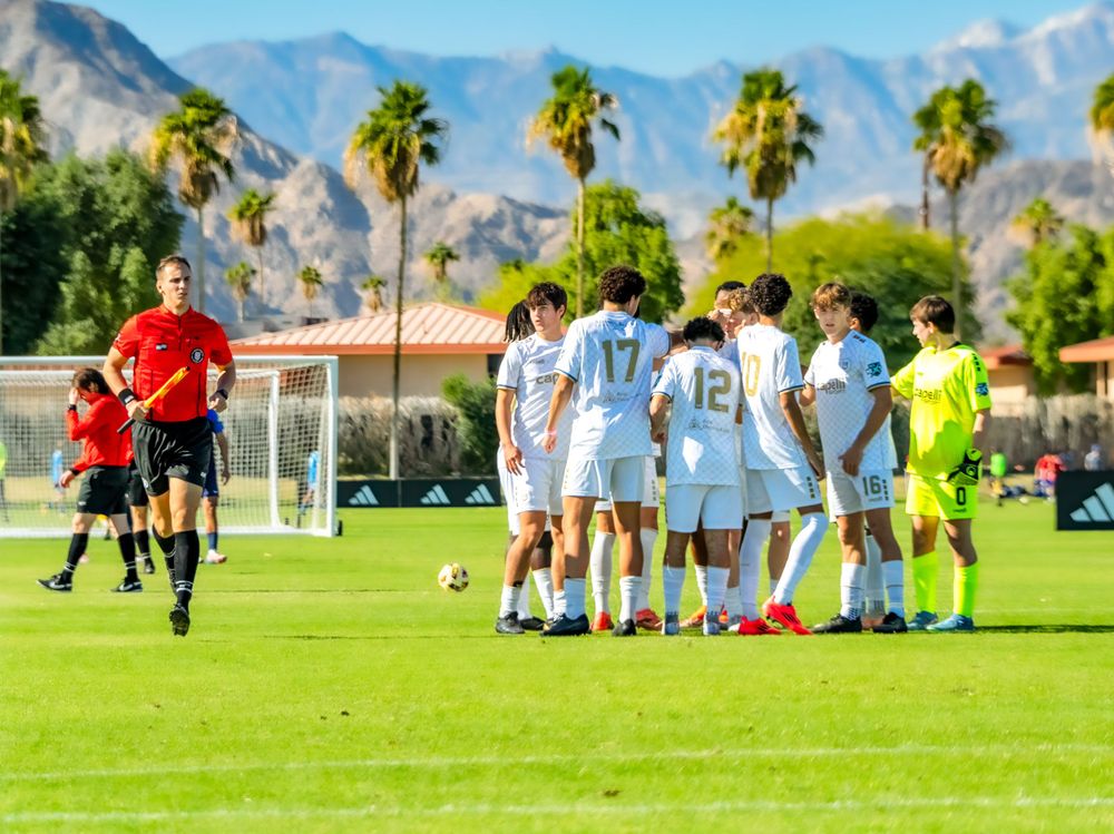 Soccer players huddle on a green field with palm trees and mountains in the background.