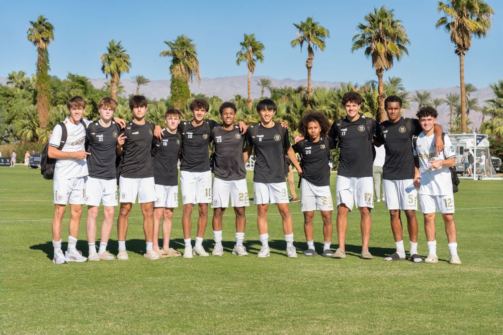 Soccer team poses for group photo on grass field with palm trees behind them.