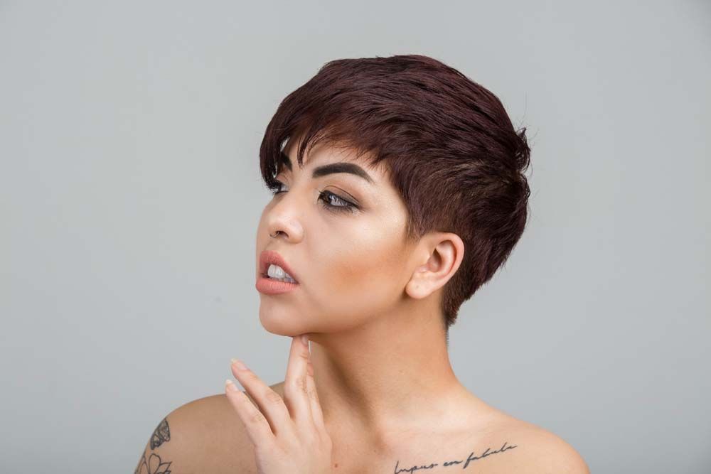 Profile view of figure with short dark pixie haircut and bare shoulders against gray background.