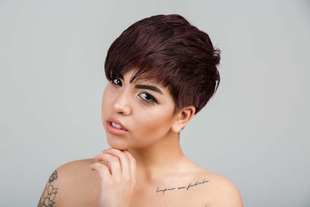 Short pixie haircut portrait with a hand on chin pose, showing script tattoo on shoulder against gray background.