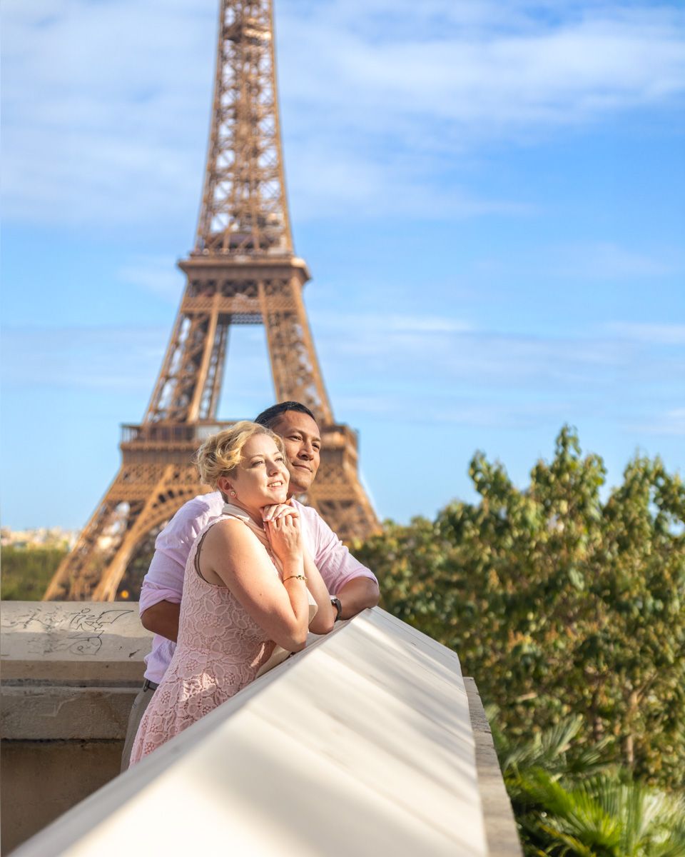 A romantic couple embraces on a terrace overlooking the Eiffel Tower against a blue sky.