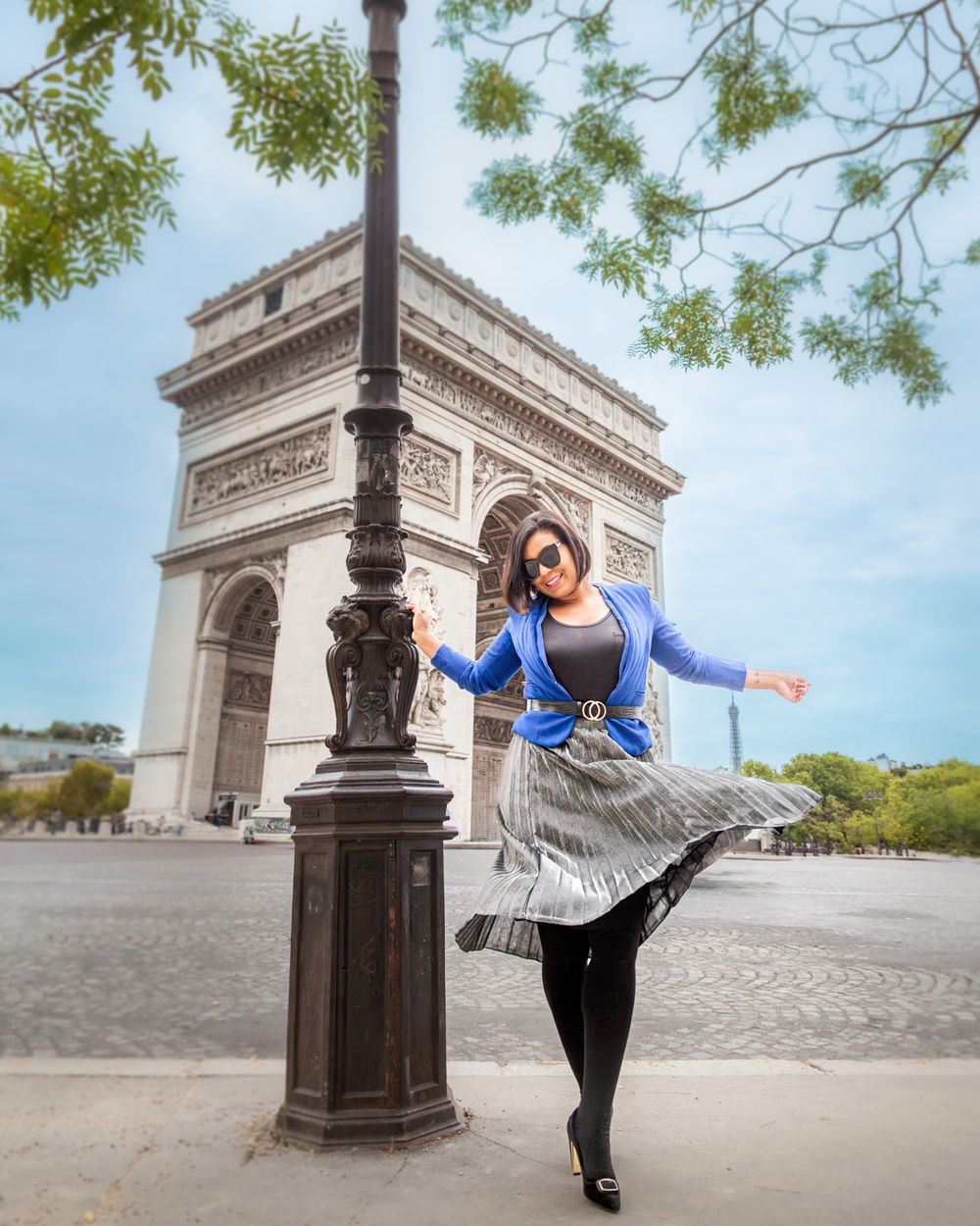 A joyful tourist in a blue jacket twirls near the iconic Arc de Triomphe monument in Paris on a sunny day.