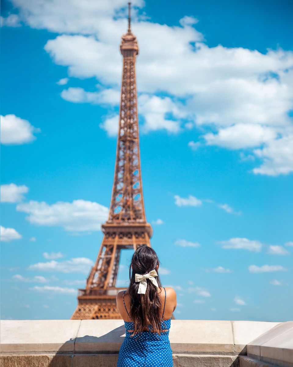 Tourist in a blue dress admires the Eiffel Tower against a bright blue sky with white clouds in Paris, France.