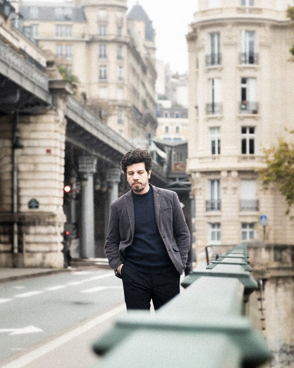 A stylish figure in a dark blazer and sweater walks along a classic Parisian street with elegant limestone buildings.
