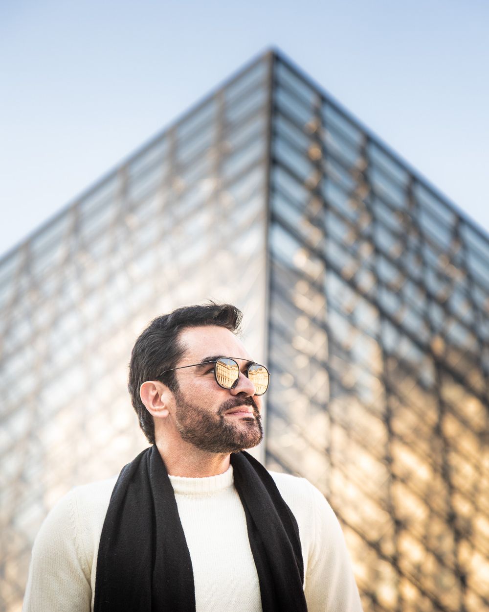 Stylishly dressed person wearing sunglasses stands in front of a modern glass building on a sunny day.