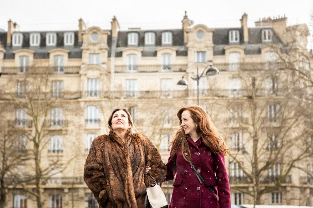 Two friends wearing winter coats stroll past elegant Parisian apartment buildings with ornate architecture.