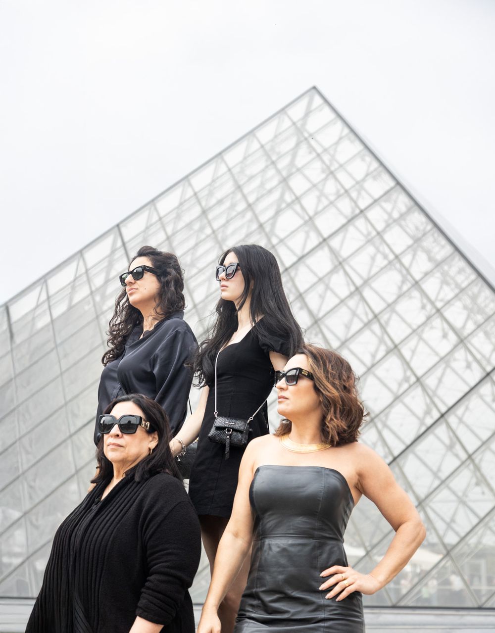 Four people in black outfits pose fashionably in front of a modern glass pyramid structure.