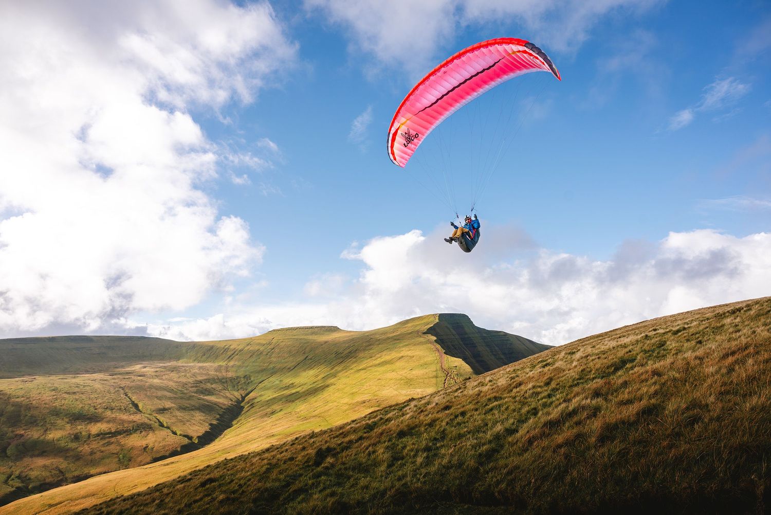 A paraglider with a pink wing soars above rolling green hills and mountains against a cloudy blue sky.